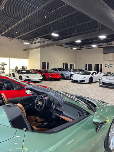 The image captures the interior of a car dealership, likely located in West Chicago, United States, given the context of the query. A line of high-end sports cars are parked in a row, with a green Ferrari prominently featured in the foreground. The interior of the Ferrari is visible, showcasing its tan leather seats and black steering wheel. Behind the Ferrari are other luxury vehicles, including several Lamborghinis in white, red, and grey. The dealership's architecture includes a high ceiling with exposed beams and industrial lighting. The walls are painted a neutral beige color, with windows and framed pictures visible in the background.