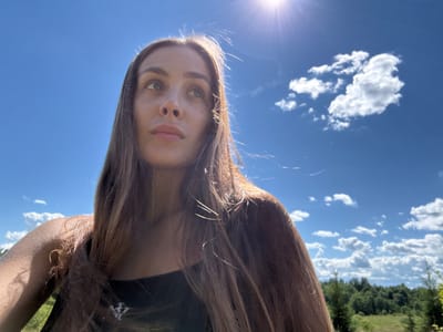 This is a close-up, low-angle, upside-down selfie of a young woman with long, light brown hair. She is outdoors, with a bright blue sky and scattered white clouds filling the background. A sun flare is visible in the lower left corner of the image. The upper portion of the image shows a blurry, inverted landscape with green trees and more clouds. The woman is wearing a dark top. She has green eyes and light skin. The lighting suggests it is daytime. The location context is Sholokovo, Russia.