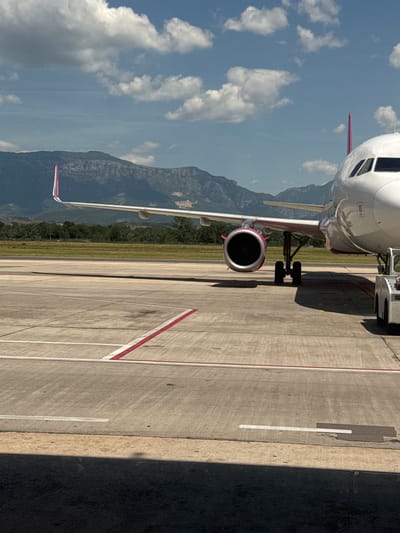 A Wizz Air Airbus A320 aircraft is parked on the tarmac at Tirana International Airport, identified by its distinctive pink wingtips and engine nacelles. The plane's nose is partially visible on the right side of the frame, with its cockpit windows facing left. The aircraft's landing gear is extended and supporting its weight on the concrete surface. The wing stretches across the mid-ground of the image, with the engine clearly visible. In the background, a line of lush green trees separates the tarmac from a range of imposing, rocky mountains under a bright blue sky dotted with white, fluffy clouds. The airport ground is marked with white and red lines, indicating taxiways or parking positions. A small service vehicle, possibly a pushback tug, is positioned near the aircraft's nose gear on the right. The scene is bathed in sunlight, casting shadows on the tarmac. The location is confirmed to be the vicinity of Kruja, Albania, as the airport serves the capital city and the surrounding region. There are no people visible in the image.