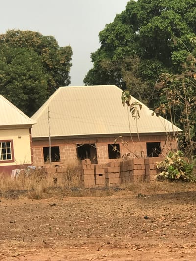 The scene, located in Egboje, Nigeria, depicts a static outdoor environment under an overcast, light grey sky. In the midground, an unfinished, single-story building constructed of reddish-brown concrete blocks is visible, featuring multiple open window and door apertures lacking frames or coverings. It is topped with a light beige corrugated metal hip roof. Directly in front of this structure, several stacks of identical reddish-brown concrete blocks are present. To the left, a portion of an adjacent, more complete building is visible, displaying cream-colored exterior walls with a maroon base trim, a white-framed multi-pane window, and a matching light beige corrugated metal roof. A white garment is visible hanging from the exterior near the base of this building. The foreground consists of dry, reddish-brown earth sparsely covered with desiccated yellowish-brown vegetation. The background is dominated by dense green foliage of mature trees. No individuals are present, and no active work is observed.