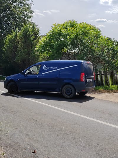 A blue delivery van with "БЕЛПОШТА" (Belpost) branding is driving on a paved road in Воўчынскі сельскі Савет, Belarus. The van has a license plate reading "AM4194-1". The van is dark blue with a white diagonal stripe and logo on the side. A person is visible driving the van. The road has a white dividing line. In the background, there are trees and a wooden fence. The sky is cloudy. The image appears to be taken during the daytime.