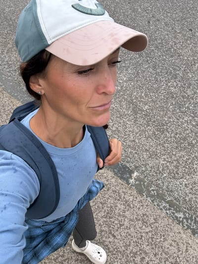 A woman, viewed from above, walks on a concrete pathway with scattered stones. She wears a blue long-sleeved shirt, a blue plaid shirt over it, black leggings, and white clogs. A grey backpack is on her shoulders. She has a baseball cap on her head, white and pink, with a greenish emblem on the front. Her hair is dark brown, and she wears a small silver earring in her visible ear. The scene is outdoors, with a grey, textured ground surface. The image does not provide any information about the location in the city of Malyi Mayak, Ukraine.