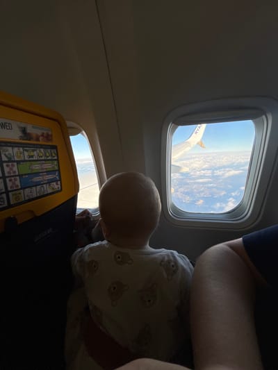 An interior view of a Ryanair aircraft cabin shows a young child, visible from behind, seated next to a window. The child has a light-colored, balding head and wears a white garment patterned with brown teddy bears. Their attention is directed towards the aircraft window on the right. Through this window, a portion of the white airplane wing, bearing the "RYANAIR" logo in blue text, is visible against a clear blue sky above a dense layer of white clouds. Below the cloud layer, distant brown landmasses, possibly mountains, are discernible. A second aircraft window to the child's left shows a similar view of the sky and clouds. To the far left, a yellow-framed seatback tray table with a printed safety instruction card is partially visible. The cabin's ceiling is light gray. A partial view of an adult's arm, wearing a dark blue or black sleeve, is present in the lower right foreground, indicating another passenger. The aircraft is in flight, likely in airspace associated with an arrival at or departure from Girona-Costa Brava Airport in Vilobí d'Onyar, Spain.