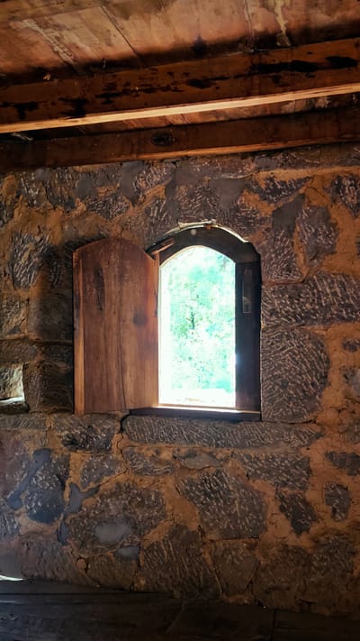 A window with a wooden shutter ajar is set in a stone wall inside a building with a wooden ceiling, likely in the Sà Phìn Commune, Vietnam. The window frame is arched. Light streams through the open portion of the window, illuminating the trees outside. The stone wall is rough-hewn and mortar appears to have been applied between the stones. The ceiling is made of roughly finished planks of wood. The floor is also wooden and dark. The shutter appears to be made of a single piece of wood and is open.
