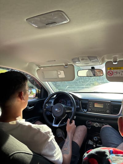 A male driver is seated in the driver's seat of a Kia vehicle. He has short, dark hair and is wearing glasses and a white shirt. His left arm has a visible tattoo. His hands are on the steering wheel. A passenger is seated in the passenger seat. The vehicle appears to be parked. The image was taken from inside the vehicle. The environment outside the car is not clearly discernible. The specific location in the city of Mihailovo, Bulgaria is not identifiable based on the provided image.