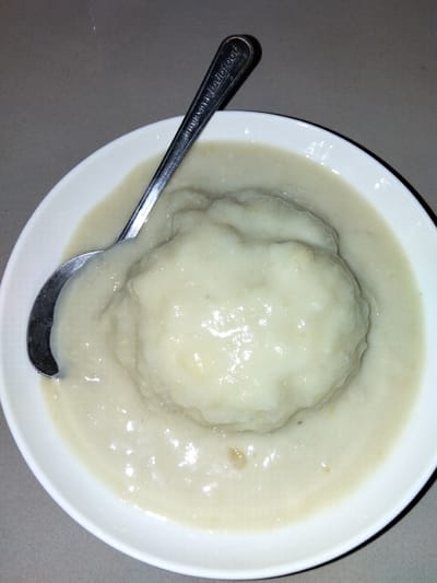 A close-up shot shows a white bowl filled with a light-colored, creamy sauce and a large, pale dumpling or ball of mashed food in the center. A silver spoon rests on top of the food, with the handle extending out of the bowl and onto a grey, textured surface. The spoon has text embossed on it, reading "Indofood". The background is blurred, showing more of the grey surface. The lighting is somewhat dim, creating highlights on the wet surface of the food and the spoon. There are no people visible in the image. This image depicts a single serving of food, likely a traditional dish, presented in a simple bowl, possibly at a restaurant or home setting. There is no information about the location in Geulanggang Teungoh, Indonesia.