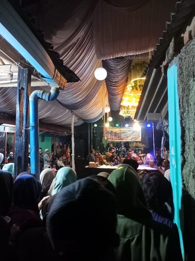 This media file captures an evening community event or performance in Cimaung, Indonesia. The image is taken from within a crowd, looking towards a brightly lit stage under a canopy of draped fabric.

In the foreground, the backs of numerous attendees are visible, many wearing head coverings like hijabs or hoodies, indicating a diverse audience gathered to watch the event. To the left, a wooden post and a bright blue gutter or pipe structure are prominent, suggesting a semi-permanent or temporary outdoor setup.

The main focus of the scene is the stage, where several individuals are seated, performing with traditional musical instruments, including drums and possibly other string or percussion instruments. Large speakers flank the stage, and bright stage lights illuminate the performers. Behind the performers, a banner is displayed. Visible text on the banner includes "BRIGHT SYAHIR" and, below it, "BERSHOLAWAT," or "BERSHOLAWAT BERSAMA...", with additional, less legible text likely detailing the event or sponsors.

Overhead, a ceiling of light-colored, draped fabric creates a tent-like atmosphere, decorated with several glowing white spherical lanterns. Further back, another section of ceiling fabric is illuminated with warm, yellow lights. To the right, a rough, corrugated roof structure and a bright teal wall are visible, framing the scene. The overall lighting suggests it is nighttime, with artificial lights providing illumination for the event. The atmosphere appears communal and engaging, centered around the musical performance.