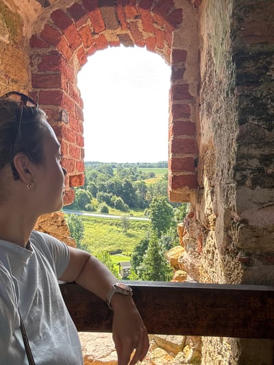 In the city of Vana-Vastseliina, Estonia, a woman is captured in a close-up shot, looking out from a window of what appears to be ancient ruins. The view through the arched window reveals a lush green landscape dotted with trees and fields under a bright sky. The woman, positioned in the foreground, is seen in profile, her head turned towards the expansive scenery. She is wearing a light blue t-shirt and a watch on her left wrist. The window frame is constructed of weathered brick and stone, showing signs of age and disrepair. A wooden railing runs horizontally across the lower portion of the image, suggesting a viewing platform or balcony. The overall impression is one of quiet contemplation, with the woman absorbed in the view of the natural beauty surrounding the historical site.