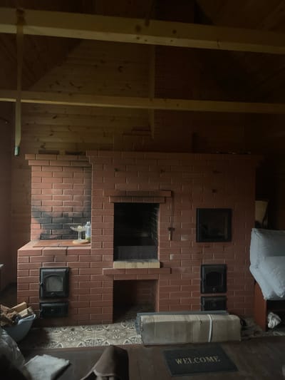 This media file displays an interior scene of a room, likely a house, in Krasnoe, Russia. The focal point of the image is a large, unfinished brick stove or fireplace that occupies a significant portion of the frame. The stove is constructed from reddish-brown bricks and features several openings, including a main firebox and what appear to be smaller access doors or vents. Some parts of the stove show signs of soot or charring, suggesting recent use or ongoing construction.
To the left of the stove, the wall is made of light-colored wooden planks, giving a rustic feel to the space. Exposed wooden beams are also visible, suggesting that this might be an attic or a room with an exposed ceiling structure. To the right of the stove, the wall features patterned tiles and a dark welcome mat with the word "WELCOME" printed on it in white. Various objects are scattered around, including stacks of boxes, a white pillow or bag, and some firewood. The lighting in the room is dim, creating a somewhat shadowed atmosphere, and it is difficult to determine the time of day or weather conditions outside. No people are visible in the image, and the overall impression is one of a work-in-progress or a utilitarian space.