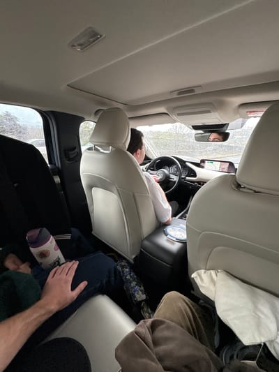 A car interior is depicted with a person in the driver's seat and individuals in the back. The driver is reclining, eyes closed, and resting their head against the headrest, wearing a white shirt. The rearview mirror reflects a person's face in the backseat. In the backseat, an individual is seated with their arm resting on their leg, wearing a dark green sweater. A patterned water bottle is visible near their leg. A white pillow or blanket is near the front passenger seat, and a brown garment is draped behind it. The car has light-colored upholstery and a sunroof. The exterior environment is visible through the car windows, with hints of foliage and buildings. The location is not directly identified as Santiago Yancuitlalpan, Mexico, in the image.