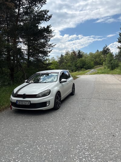 A white Volkswagen Golf, likely a GTI model, is the primary subject of the image, parked on or beside an asphalt road in what appears to be a natural, forested setting. The car is angled towards the viewer, showcasing its front end and left side. It features a black stripe running over the hood and possibly the roof, distinctive multi-spoke wheels, and a red stripe on its front grille. A red tow hook or strap is visible on the lower front bumper. On the top of the windshield, a black decal with "GOLF" in white letters is present.
The scene is set outdoors on a bright day, indicated by the clear blue sky with scattered white, wispy clouds. Lush green trees and dense foliage line both sides of the road, suggesting a rural or semi-rural environment. The road itself is made of dark asphalt with visible lighter aggregate, and it appears to curve gently into the distance.
The visible license plate on the car reads "CB 5016 CX". Below this, smaller text includes "BG" (indicating Bulgaria), and what appears to be "Umwelt Auto" or similar followed by a phone number "0877 780 333". No people are visible in the image, and there is no clear activity occurring other than the static presence of the car. The overall impression is one of a calm, scenic roadside stop in Gorna Vasilitsa, Bulgaria.