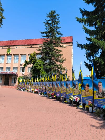 An outdoor scene in Illintsi, Ukraine, displays a memorial installation positioned along a paved area in front of a civic building identified as the "МІСЬКА РАДА" (City Council). The two-story, light-brown building features a red-tiled roof, multiple white-framed windows, and a yellow and green crest on its facade. A large evergreen tree stands centrally before the building, with additional trees framing the view. The memorial consists of a curved line of vertical banners, each approximately human height, displaying a portrait of an individual, primarily in military uniform, against a blue and yellow background with accompanying Ukrainian text. Small Ukrainian flags are mounted on poles at the top of each banner. At the base of each banner, on the red brick paving stones, numerous floral tributes, including bouquets of fresh and artificial flowers in various colors and potted plants, are arranged in dark containers. The sky is clear blue. No individuals are present in the image.