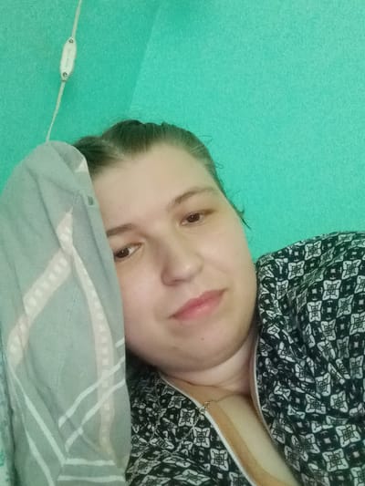 A woman is lying down with her head resting on a pillow, and part of her face is covered by a light blue and white striped blanket. She is wearing a black and white patterned shirt and a gold necklace. The background is a solid turquoise wall. A white wire with a small oval white object attached to it is visible in the upper left corner. This image was taken indoors, possibly in a bedroom or living area, in Kitskany, Moldova.