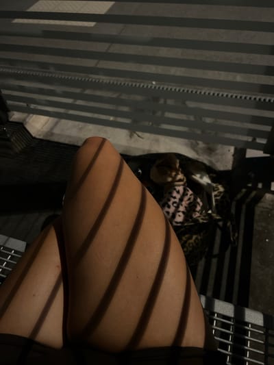 The image shows a close-up of a person's legs, with strong diagonal shadows cast across them, suggesting they are outdoors under a striped structure like a fence or pergola. In the background, partially obscured by shadows and a fence, is a dog wearing a patterned blanket. The ground appears to be concrete or pavement. The setting is consistent with an outdoor seating area in a public space. The image does not provide any specific visual cues to pinpoint the location to Ohlstadt, Germany.