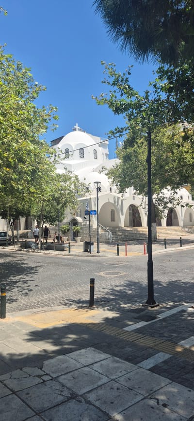 A white church with multiple domes and arched entrances is situated in a city square in Paleo Faliro, Greece. The church is surrounded by lush green trees, providing shade. The foreground features a cobblestone street with bollards and yellow tactile paving. Two people and a dog are visible near the entrance of the church, suggesting a public space. The sky is clear and blue, indicating a sunny day. The overall scene depicts a typical urban environment with a prominent religious building.