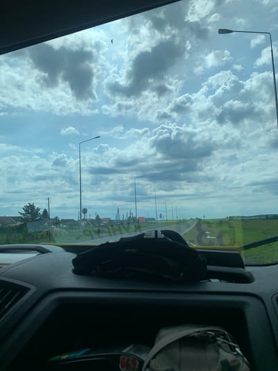 The image is taken from inside a vehicle, looking forward through the windshield. The sky is filled with dramatic, heavy clouds, with patches of bright blue sky visible. Sunlight filters through the clouds, creating a bright, almost ethereal light. A single street lamp is visible in the upper right quadrant of the image. In the distance, a road stretches out, lined with more street lamps. To the left of the road, there are houses with red roofs and some trees, suggesting a rural or semi-rural setting. The foreground of the image shows the dashboard of the vehicle, with a black cap or bag placed on it. A faint reflection of yellow signage is visible on the windshield. There is a small dark object in the sky above the clouds, possibly a bird or an insect. The overall impression is of a journey on a cloudy but bright day, passing through a village or town. The location is identified as Shlyapniki, Russia.