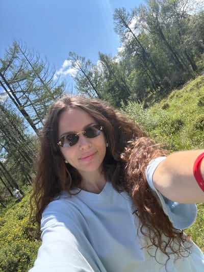A woman is taking a selfie in a wooded area with a grassy slope covered in small yellow flowers. The woman has long, curly brown hair and is wearing sunglasses, a light blue t-shirt, and a red bracelet on her right wrist. The background shows tall, slender trees, some with sparse needles, and a bright blue sky with scattered clouds. In the distance, on the grassy slope, a person in a white outfit can be seen. The overall scene is bright and appears to be outdoors on a sunny day. The location is identified as Bichiktu-Boom, Russia.