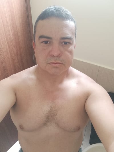 An adult male is depicted from the upper torso upwards, centered in the frame, in an indoor setting within La Merced, Ecuador. He is bare-chested, facing directly towards the camera with a neutral expression. His hair is short, dark, and shows signs of graying. He has a small silver stud piercing in his left nostril. Visible stubble covers his chin and upper lip area. His chest displays moderate dark hair concentrated centrally. The individual exhibits a light skin tone with some redness observed on his upper chest and around his nose and under his eyes. To the left of the subject is a light brown wooden door or panel. To the right is a plain, light beige wall, with a section of light-colored tiles below it. A portion of a white ceramic basin or sink and a dark, indistinct object are visible in the lower right background. The lighting is even, suggesting an indoor environment like a bathroom or private room.