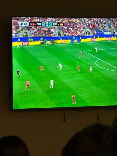 The image shows a large television screen displaying a soccer match. The score graphic at the top left of the screen indicates the time as 49:02 and the score as Portugal 0, Spain 0. The game appears to be in progress with several players on the green field. Some spectators are visible in the stands behind the field. The scene is illuminated by ambient room lighting. The television is mounted on a wall in a dimly lit room, with the lower portion of the image showing the silhouettes of two people watching the screen. The city of El Escorial, Spain, is not depicted in this image, as the image is a close-up of a television screen displaying a soccer game.
