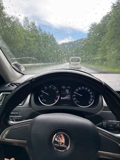 The image captures the interior of a car, looking out through a rain-streaked windshield. The driver's perspective is evident, with the steering wheel prominently displayed in the foreground. The dashboard features two main circular gauges, likely a speedometer and a tachometer, with a smaller digital display between them showing "90" and other unreadable data. The steering wheel has a Skoda emblem in the center. To the right of the steering wheel, a turn signal stalk is visible. Outside the car, the road is wet, and trees line both sides, suggesting a rural or forested area. A white van is visible ahead on the road, and the sky appears overcast with patches of blue. The scene conveys a sense of driving in the rain. The location is specified as Brishbash, Russia.