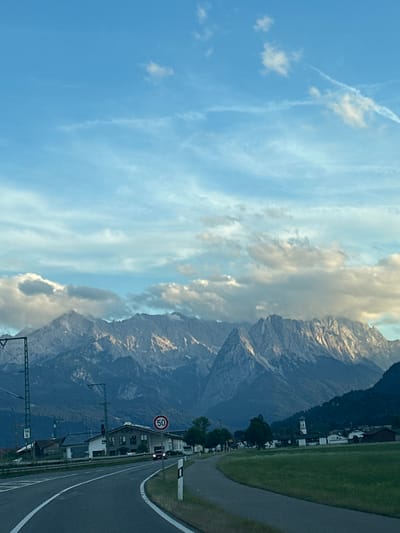 The image depicts a scenic view of mountains from a road, with a small town or village in the foreground. The sky is blue with scattered clouds, and the sun is casting light on the mountain peaks. In the foreground, there is a road with white lines marking the lanes. To the right of the road, there is a grassy field. To the left of the road, there are some buildings, traffic lights, and utility poles. A red speed limit sign indicating 50 is visible. In the distance, the town has several buildings, a church steeple, and some trees. The overall impression is that of a peaceful rural landscape with mountainous backdrop, likely taken during the daytime. The scene is consistent with views in the vicinity of Farchant, Germany, given the characteristic landscape and architectural styles.
