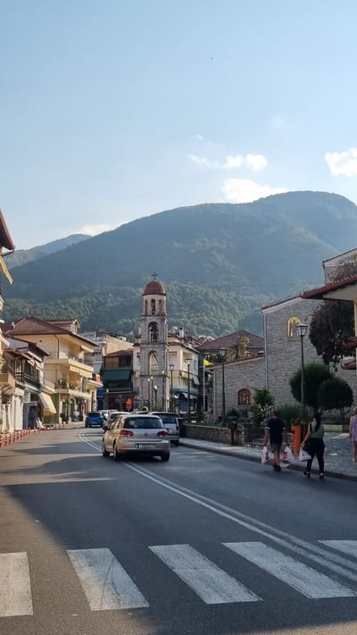 In Litochoro, Greece, a sunlit street in a town is captured. Buildings with red-tiled roofs line the street, with a prominent church tower featuring a red dome rising in the background. The tower has an arched opening with a bell and a small balcony. A gentle slope ascends towards a verdant, tree-covered mountain range that dominates the skyline under a clear blue sky with scattered clouds. Traffic moves along the asphalt road, marked with white lines and a pedestrian crossing in the foreground. A silver Volkswagen Golf is visible driving away, its license plate reading "IOH 3822." Two individuals, one carrying white shopping bags, walk on the sidewalk to the right of the street. The scene is bathed in warm sunlight, casting shadows across the road and sidewalks, indicating it is likely late afternoon.
