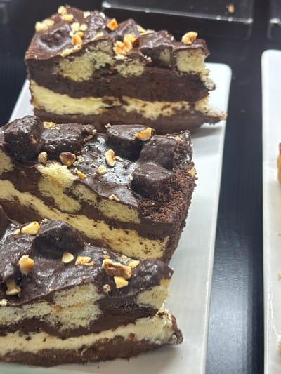 A close-up shot shows three slices of marble cake on a white serving dish. The cake has alternating layers of light and dark batter and is topped with chocolate frosting and chopped nuts. A dark, undefined surface appears beneath the dish. The location is Lipomo, Italy.