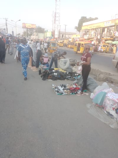 The image displays a bustling street market scene on a paved road in Ikotun, Nigeria, under bright, hazy daylight. In the foreground and midground, multiple vendors operate directly on the asphalt. Piles of assorted shoes, sandals, and bundled clothing are spread out on mats or tarps. A man in a blue and white patterned traditional outfit walks towards the viewer on the left. Center-right, a woman wearing sunglasses, a maroon "AERO GIRL" t-shirt, and black pants stands, holding items, with a light-colored hat hanging from her arm. Behind her, a man in a plaid shirt is bent over a large pile of goods next to a white plastic bag, while a person in a pink top sits on the ground nearby. The background reveals numerous pedestrians, several yellow Keke Napep (auto-rickshaws) on the road, and commercial buildings with storefronts, including one marked "DE WIC MARKET." Overhead utility poles, power lines, and prominent red and white telecommunications towers are visible against the sky. A light-colored vehicle is partially visible on the far right.