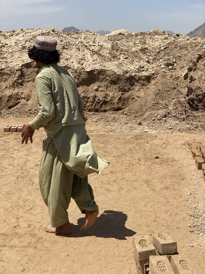 A man wearing a light green shalwar kameez and a patterned hat walks barefoot across a dusty ground in Deh-E Bagh, Afghanistan. A large mound of dirt and earth is behind him, and mountains are visible in the distance. Stacks of unbaked bricks are arranged on the ground.
