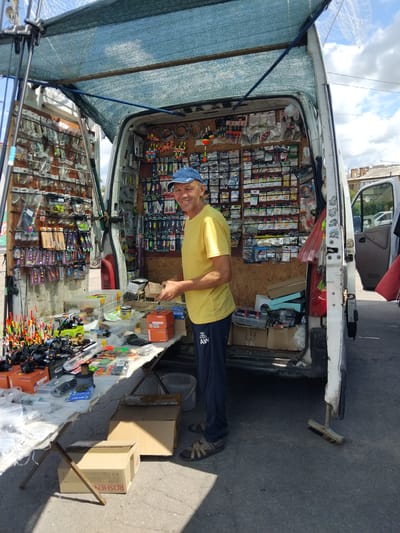 A man in a yellow t-shirt and a blue baseball cap is standing in front of an open-backed white van, which is acting as a stall selling fishing gear. The van's interior is packed with fishing lures, floats, lines, and other accessories, organized on shelves and pegboards. The man is holding a small box and appears to be looking at the camera with a smile. In front of the van, a table displays more fishing equipment, including fishing rods and reels. The scene is outdoors, with a street or paved area in the foreground and buildings visible in the background, suggesting an urban or semi-urban setting. The sky is partially cloudy. The overall impression is a market stall or roadside vendor.
