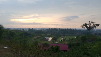 The image depicts a landscape in Uruekanoe, Indonesia, at sunset or sunrise. In the foreground, lush green vegetation dominates with a building having a red roof partially obscured by trees. Further back, a small pond with a structure stands out amidst the greenery. Beyond the pond, a dense line of trees stretches across the midground. In the distance, communication towers and a mosque are visible on the horizon. The sky is filled with soft, muted colors of the setting or rising sun, interspersed with clouds. A large tree stands prominently on a raised area to the right. A patch of waste material is visible on the ground in the lower left.
