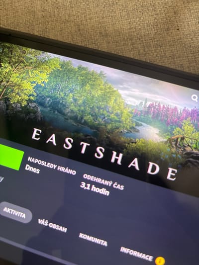 This is a close-up, angled shot of a digital screen, likely a computer or gaming console, displaying a video game interface. The game's title, "EASTSHADE," is prominently displayed in large, white, sans-serif letters across the screen. Beneath the title, there is information about game activity, including "NAPOSLEDY HRÁNO" (Last Played) and "Dnes" (Today), indicating recent engagement. "ODEHRANÝ ČAS" (Time Played) is shown as "3,1 hodin" (3.1 hours).
The background of the game appears to be a lush, vibrant landscape. A body of water reflects a sky that is partially visible, with hints of clouds. The landscape is populated with trees, some bearing clusters of bright purple or pink blossoms, while others are laden with bright green foliage. The overall impression of the game's visual style is one of fantasy and natural beauty.
On the right side of the screen, there are additional menu options in white text: "KOMUNITA" (Community), "VÁŠ OBSAH" (Your Content), and "INFORMACE" (Information), the last of which is accompanied by an exclamation mark icon within a yellow circle, often used to denote alerts or important notices. At the bottom, a button labeled "AKTIVITA" (Activity) is visible, highlighted in a light grey, rounded rectangular shape. The screen is placed on a textured, light-colored surface, possibly a rug or fabric. The camera angle and lighting suggest the photo was taken indoors. There is no indication of people or a specific time of day or weather within the frame itself, other than what is depicted in the game's artwork.