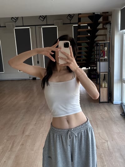 A young woman with dark hair is taking a selfie in a well-lit room. She is wearing a white tank top and grey sweatpants. Her hair is long and dark, falling over her shoulders. She is holding a light-colored smartphone in her right hand, capturing her reflection in a mirror. Her left hand is raised, and she appears to be adjusting her hair or posing.
The background shows a modern interior with a wooden floor. To the left, a reflection reveals a staircase with black metal railings and wooden steps. There are shelves with various items on them, including what appear to be bottles and decorative objects. A white ceiling with recessed lighting is visible. The overall impression is of a home environment, likely a living room or a dressing area. The lighting suggests it is daytime. There is no visible text in the image.