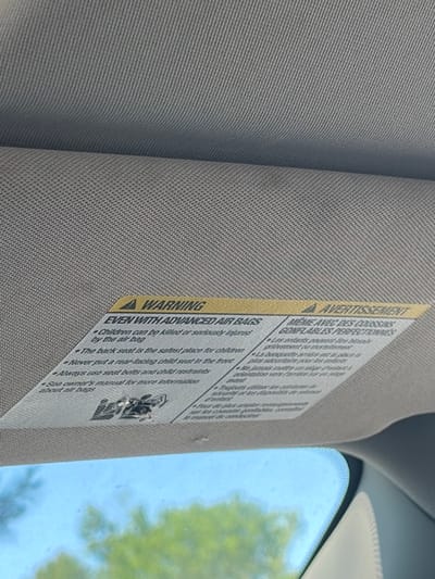 The image provides a close-up view of the interior of a car, focusing on a beige or light gray sun visor. The sun visor is made of a textured, woven fabric. Attached to its surface is a prominent yellow and white safety warning label, positioned vertically.
Through the car's windshield, visible in the background, a bright, clear blue sky and blurred green foliage from trees are apparent, indicating an outdoor, daytime setting with sunny weather. The car's headliner and a portion of the windshield's frame are also visible, bordering the exterior view.
The warning label contains bilingual text in English and French, cautioning about airbag safety, particularly concerning children. The English section, under the heading "A WARNING EVEN WITH ADVANCED AIR BAGS," states: "Children can be killed or seriously injured by the air bag," "The back seat is the safest place for children," "Never put a rear-facing child seat in the front," "Always use seat belts and child restraints," and "See owner's manual for more information about air bags." A small graphic depicting a child and an airbag is present near the bottom of the English text. The French section, titled "AVERTISSEMENT MÊME AVEC DES COUSSINS GONFLABLES PERFECTION," provides similar safety guidelines.