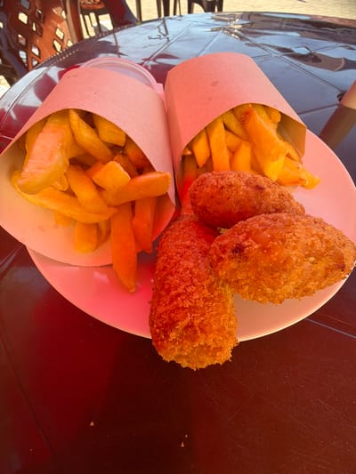 The image displays a plate of food on a dark, reflective surface, likely a table. On the plate are two paper cones filled with golden-brown french fries. Next to the fries are three crispy, breaded croquettes. The food appears to be served outdoors, with a hint of a checkered pattern on the reflective surface, possibly from a tabletop. The lighting suggests it's daytime, with some shadows indicating the presence of overhead structures or furniture. There are no people visible in the image. The overall presentation suggests a casual dining setting. The context of Amzara, Abkhazia is not visually discernible from the provided image.