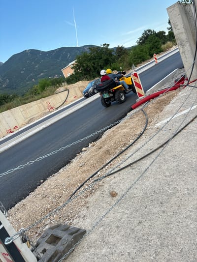 The image depicts a construction site in Radanovići, Montenegro, with a new asphalt road being laid. In the foreground, a gray concrete surface is visible with a black traffic barrier, a chain, and a padlock. The road itself is paved with dark asphalt and bordered by a white line. Construction materials, including red corrugated pipes and gravel, are present along the side of the road. A yellow quad bike carrying two people, one wearing a white helmet and the other a yellow helmet, is driving on the asphalt. Behind the quad bike, a dark car is also on the road. The background features a lush green mountain slope with trees and a building, under a clear blue sky with a contrail. The overall scene suggests ongoing road construction and activity in a scenic, mountainous region.