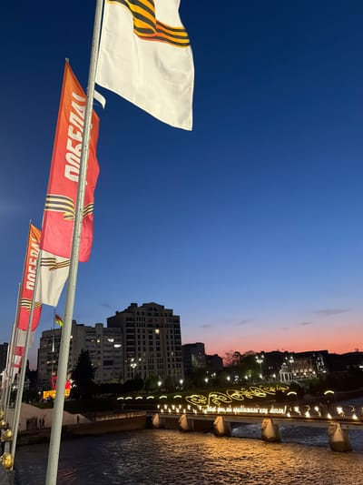 The photo, taken in the city of Chermen, Russia, shows a row of celebratory flags fluttering in the foreground against a backdrop of buildings illuminated with evening lights. The flags feature the St. George ribbon, a symbol associated with Victory Day, and the Russian word "ПОБЕДА" (POBEDA), meaning "Victory".
The sky transitions from a deep blue to a soft pink-orange hue near the horizon. A bridge, adorned with decorative lights and script in Russian that translates to "This day, we brought as close as we could!", spans a body of water, reflecting the bridge's illuminations.
The urban environment consists of several multi-story buildings with lit windows, indicating residential or commercial activity. The image is taken from a slightly low angle, emphasizing the flags and the depth of the cityscape.