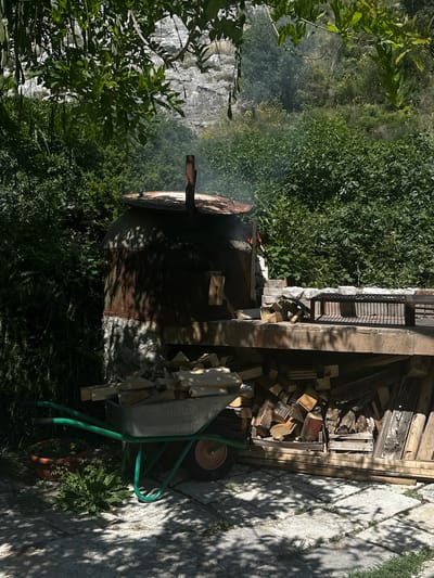 This outdoor scene in Bartula, Montenegro, features a rustic cooking or heating installation set against a lush, vegetated hillside under bright sunlight.
In the center-left, a large, dark, cylindrical metal structure, exhibiting significant rust and held together by rivets or seams, serves as an oven or smoker. It rests on a rough-hewn stone base and has a rusty metal chimney pipe protruding from its slightly conical top. Smoke is visibly rising from the top opening. Dappled shadows from overhead foliage pattern its surface and the stone base.
To the right of the oven, a sturdy, light-colored stone or brick counter provides a workspace. On this counter, a rectangular black metal grill grate is positioned. Smaller pieces of wood are scattered on the counter surface near the oven. Beneath the counter, a substantial quantity of cut firewood and various pieces of lumber are neatly stacked.
In the lower-left foreground, a bright green metal wheelbarrow, equipped with a single orange wheel, is positioned on a paved surface. It contains a load of split firewood. Adjacent to the wheelbarrow, a brown clay pot holds a small plant. The ground in the foreground is paved with light-colored, rectangular stone slabs, with grass visible in the gaps, and covered by strong, irregular shadows cast by overhead tree leaves.
The background is dominated by dense green foliage, comprising various bushes and trees. Beyond the immediate vegetation, a large, light-grey, rocky cliff face or steep hillside is partially visible through the trees. The scene is illuminated by strong natural sunlight, creating high contrast and distinct shadows. No people are present in the image.