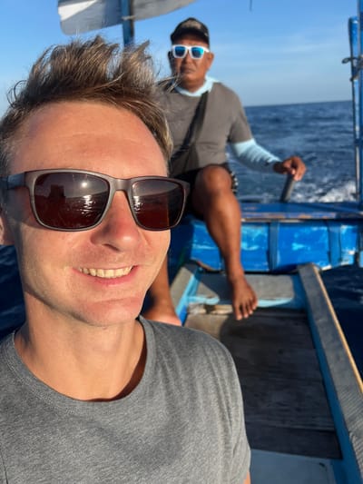 A selfie photograph taken on a boat. A light-skinned man with short brown hair, wearing sunglasses and a gray t-shirt, smiles at the camera. In the background, a dark-skinned man wearing sunglasses and a gray shirt with light blue sleeves rows the boat with a paddle. The boat is blue. The setting appears to be the sea. The photo was taken in Seririt, Indonesia.