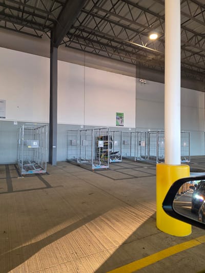 The media file shows a scene inside a large industrial building, possibly a warehouse or distribution center, located in Roseville, USA. The perspective is from inside a vehicle, as evidenced by the side mirror reflecting a car and a portion of the vehicle's interior. The main subjects are several metal cage trolleys lined up in a row. These trolleys are commonly used for transporting goods.
The building's interior features a high, industrial ceiling with exposed metal beams and a large white pipe running horizontally across the frame. Lighting is provided by overhead industrial lamps, one of which is clearly visible. The walls are painted in a neutral palette of gray and white. The floor is concrete, marked with yellow lines and dark grid patterns, suggesting designated pathways or storage areas.
The time of day appears to be late afternoon or early evening, as indicated by the long shadows cast across the concrete floor, illuminated by sunlight entering from an unseen source. The weather is not directly discernible, but the interior setting implies it's not an outdoor scene.
There is some visible text: a poster on the wall with green and white graphics and text, and labels on the trolleys, though the text on the trolleys is too small to read clearly. One poster seems to have the word "TABLE" printed on it near the bottom edge of the frame.
The overall impression is one of an organized, functional industrial environment. No people are visible in the frame. The scene conveys a sense of quiet operation, characteristic of a storage or logistics facility.