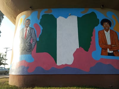 A mural under a bridge or overpass in Tawpo, Nigeria, features a painted map of Nigeria flanked by portraits of two individuals. The map is rendered in the colors of the Nigerian flag: green, white, and green. To the left of the map is a man with glasses wearing a grey suit and red tie. To the right of the map is a woman with glasses wearing a brown blazer and white shirt. The background of the mural is painted in shades of blue, pink, and purple. The base of the wall is brown, and green grass is visible at the very bottom. A car and lamppost are partially visible in the background.