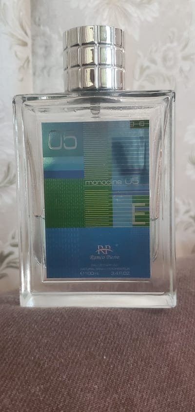 A close-up, eye-level, indoor shot shows a clear glass bottle of perfume, likely sitting on a brown fabric surface. The bottle has a silver, ridged cap and a label featuring abstract geometric patterns in shades of green and blue. The label reads "monocline 05" in white text, with the brand "Ramco Pierre" and "EAU DE PARFUM" also visible, along with "e 100ml 3.4 FL.OZ." The background appears to be a light-colored, patterned wallpaper with a floral motif. The lighting is soft and diffused. The image does not contain any discernible information about the city of Konakovo, Russia.