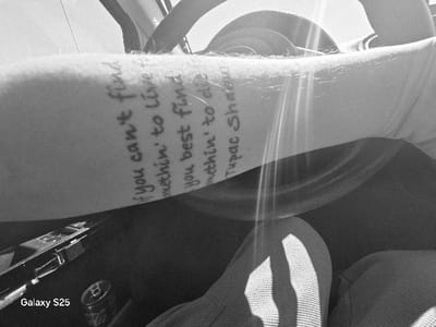 An monochromatic image depicts a first-person view inside a vehicle, located in Novo Delchevo, Bulgaria. The central subject is a forearm, oriented diagonally from the lower left towards the upper right, bearing a tattooed quote in handwritten style: "If you can't find somethin' to live for you best find somethin' to die for. Tupac Shakur." Visible below the forearm are parts of the individual's lap and legs, covered in light-colored, textured fabric. To the left of the lap area, a silver Coca-Cola can is present, partially obscured. Directly below the can, a dark electronic device, identified by the text "Galaxy S25," is visible. A portion of a dark steering wheel is visible in the upper right quadrant of the frame. Strong sunlight enters from the upper right, creating lens flares and bright highlights across the arm and steering wheel, casting shadows elsewhere within the vehicle's dark interior. No other individuals or direct interactions are visible.