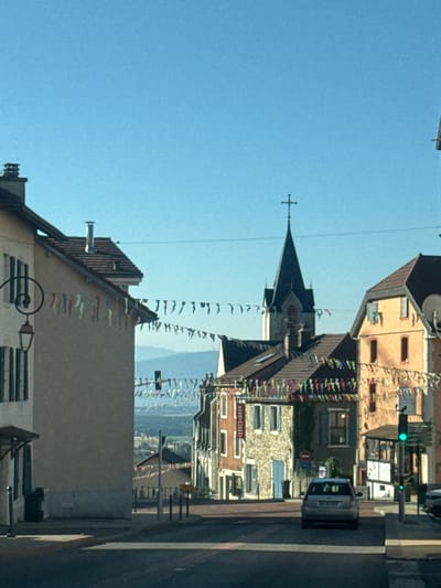 The image shows a street scene in the city of Gex, France. The view is directed towards a town with buildings and a church spire with a cross on top. Colorful pennant banners are strung across the street, suggesting a festive occasion. In the foreground, a silver car is stopped at an intersection, with a traffic light displaying a green signal. The environment appears to be a clear day with a bright blue sky and distant hills visible in the background. The architecture of the buildings suggests a European town.