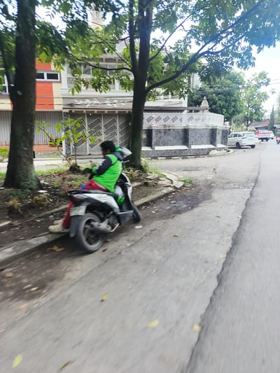 A person sits on a parked scooter on the side of a road. The person wears a bright green jacket, red pants, and white shoes. The scooter is white and black. Trees flank the road on both sides, and a building is partially visible behind the trees. In the distance, cars are parked along the street. The road is gray, and the sky is overcast.