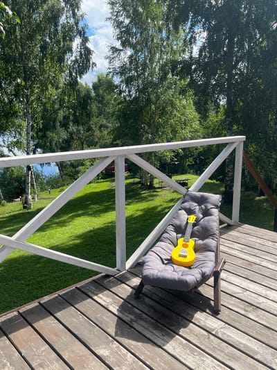 This image shows an outdoor wooden deck with a gray lounge chair placed on it. A bright yellow ukulele rests on the cushion of the chair. The deck overlooks a lush green lawn with trees in the background. A white railing separates the deck from the lawn. Beyond the lawn, a body of water is visible, and more trees frame the scene. The sky above is partly cloudy. The location appears to be a serene natural setting, possibly a backyard or a recreational area in Naberezhnaya, Russia, given the context. There are no people visible in the image.