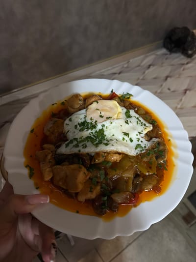 The image shows a close-up, top-down view of a white plate filled with a meal. The meal consists of cubed chicken, vegetables such as peppers and mushrooms, and a fried egg served on top. Chopped parsley is sprinkled over the dish. The plate has a decorative, wavy edge.

The plate is placed on a surface that appears to be a tiled countertop, with a metallic edge visible. In the background, a dark, lumpy object is visible on the left side, possibly trash or a bag. The lighting suggests an indoor setting, possibly a kitchen or dining area. A person's hand with manicured nails is holding the edge of the plate on the right side of the image. There is no visible text in the image. The overall impression is of a home-cooked meal, ready to be eaten.