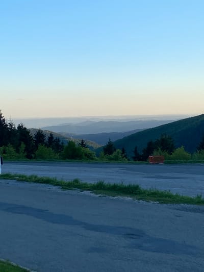The image shows a view from a road or paved area in Loučná nad Desnou, Czechia. In the distance, there are layered mountain ranges under a gradient sky of light blue to pale yellow/white. Along the roadside, there are trees and grassy areas. The road surface appears damp or wet. A bench is slightly visible amidst the trees to the right.