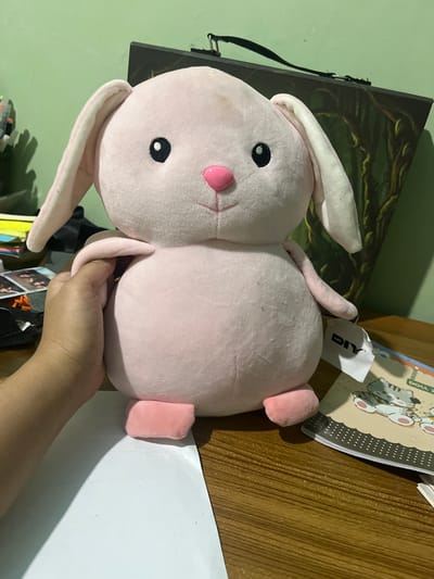 A plush pink bunny toy is being held by a person's hand, lying on a wooden surface. The bunny has black button eyes, a pink nose, and long, floppy ears. The background is a green wall and a wooden desk. On the desk, there is a notebook with cartoon cat illustrations and a white tag with the word "DIY" visible. The scene appears to be indoors, likely in a home or office setting, and there are no indications of the time of day or weather. The overall impression is casual and domestic.