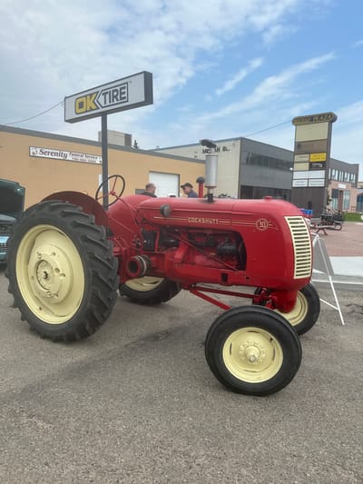 This media file is a vertically oriented, medium-angle shot of a bright red vintage Cockshutt 30 tractor parked on an asphalt surface. The tractor, a prominent object, has yellow rims on its large, black tires and a white and gold grille. Visible text on the tractor includes "COCKSHUTT" and a circular emblem with "30" inside.
The scene is set outdoors, likely during daytime with a cloudy sky. In the background, several buildings are visible. One building has a sign that reads "MICHAN PLAZA" with various business names listed below it, including "AMR," "SVENSEN'S HOLDINGS LTD," and "Century21." Another sign, clearly visible in the foreground, displays "OK TIRE" in large, bold yellow letters. Adjacent to it is a sign for "Serenity Funeral Service" with a phone number.
Several people are present, though their faces are not clearly discernible. One person appears to be looking at the tractor, and another is partially visible near the steering wheel. Bicycles are parked in the background near the buildings.
The overall impression is that of a community event or display, possibly a tractor show or a local gathering, given the presence of vintage machinery and business signage. The weather appears to be overcast. The location is identified by the "OK TIRE" and "MICHAN PLAZA" signs, and the context mentions the City of Leduc, Canada.